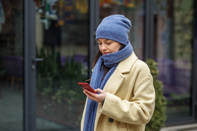 Elegant woman in knitted hat and scarf in beige coat sending text message using smartphone outside in city in winter.