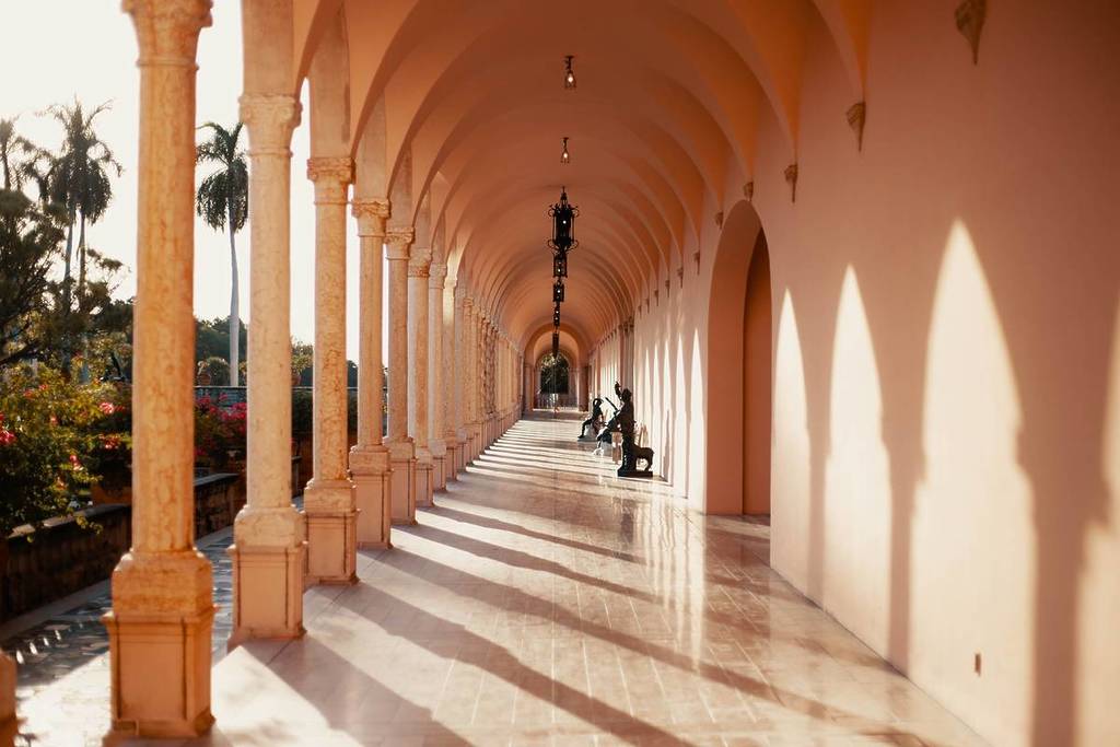 Un pasillo bellamente iluminado por el sol, con arcos y estatuas que proyectan sombras, que muestra toda la elegancia arquitectónica.