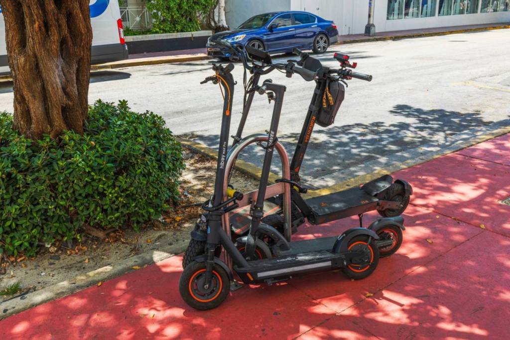 Close up view of electric scooters parked on red sidewalk near tree and cars on Collins Avenue. Miami Beach. USA. 04.15.2025.