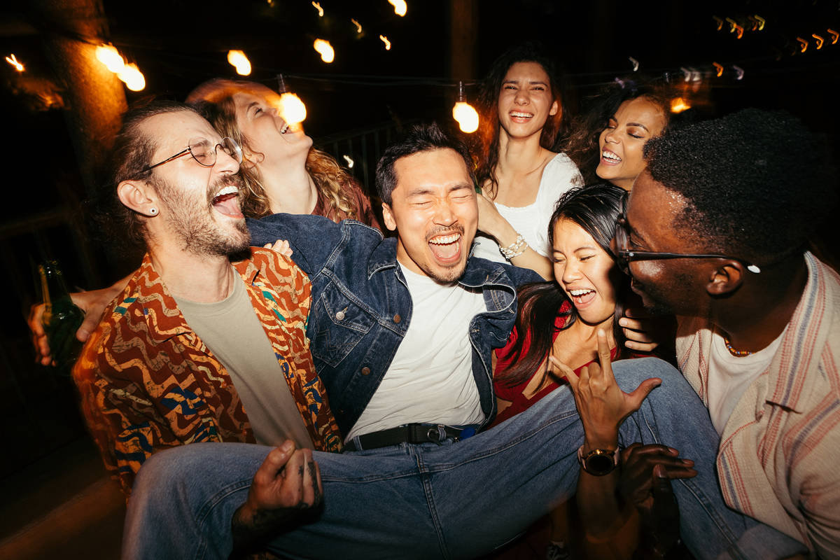 A multicultural group of friends are lifting their male tipsy friend at the rooftop night outdoor party.