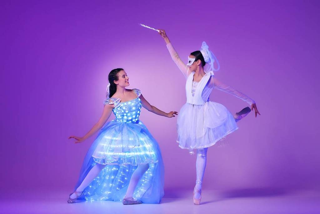 Zwei Ballerinas tanzen bei Ballet of Lights: Cinderella in funkelnden Kostümen. Die rechte Tänzerin trägt eine Maske und hält einen Zauberstab über die linke Tänzerin.