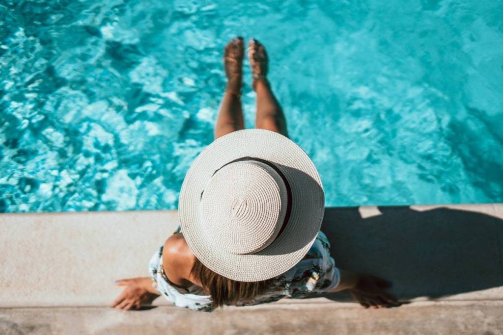 A woman wearing a hat sits at the edge of a swimming pool with her feet in the water