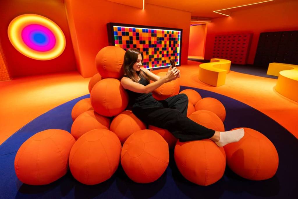 Frau sitzt bei "Dream Rooms" auf einem großen Bällehaufen und macht ein Selfie, weitere Traum-Installationen im Hintergrund