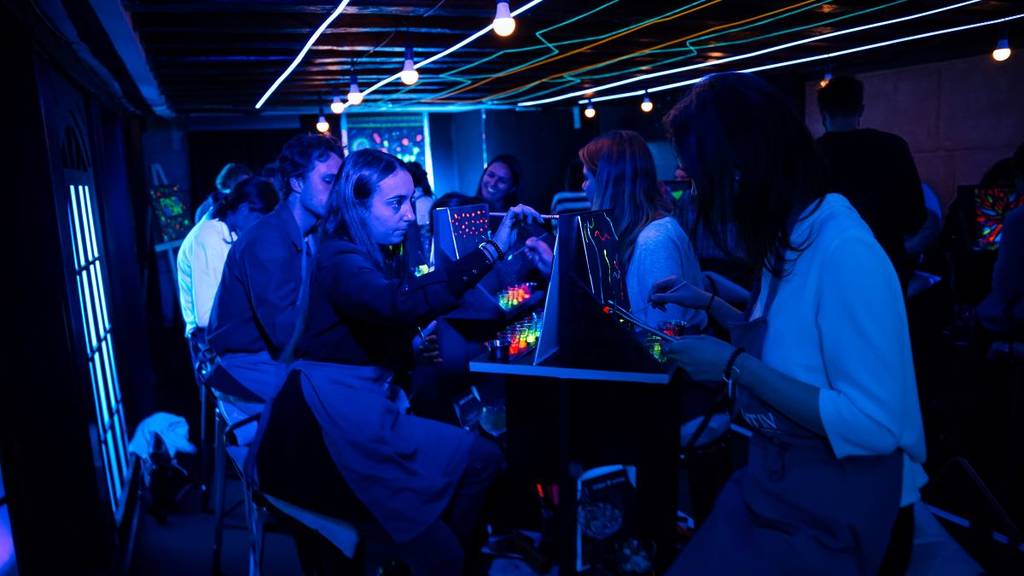 Attendees sitting up at a high table and painting in a neon-lit setting at Neon Brush.