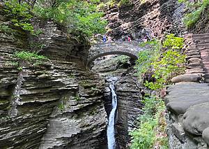 a waterfall flowing down in the middle of a gorge with green surrounding it and a stone bridge in the background