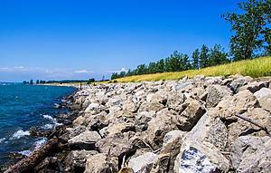 Exterior daytime stock photograph of rocks lining the shore of Lake Erie with grass and trees on side against blue sky backdrop in Buffalo, New York in Erie County in summer