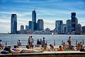 New York City, USA - June 09, 2019: A large group of people are seen relaxing on a warm summer weekend afternoon on a pier of  Hudson River Park  in Lower Manhattan. Behind them, across the Hudson River is a cityscape of skyscrapers in Jersey City, New Jersey.