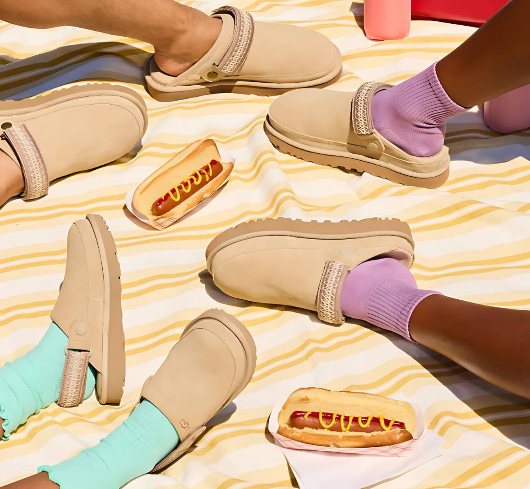 People's feet in brightly colored socks and UGG clogs on a picnic rug with hot dogs.