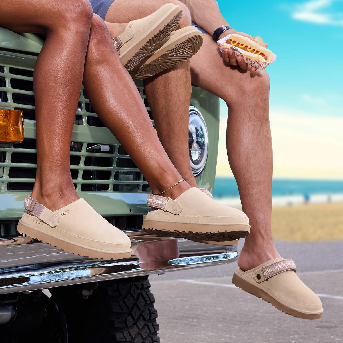 Several people's legs draped over the front of a car. They are wearing UGG Mustard Seed clogs and one of them is holding a hot dog.