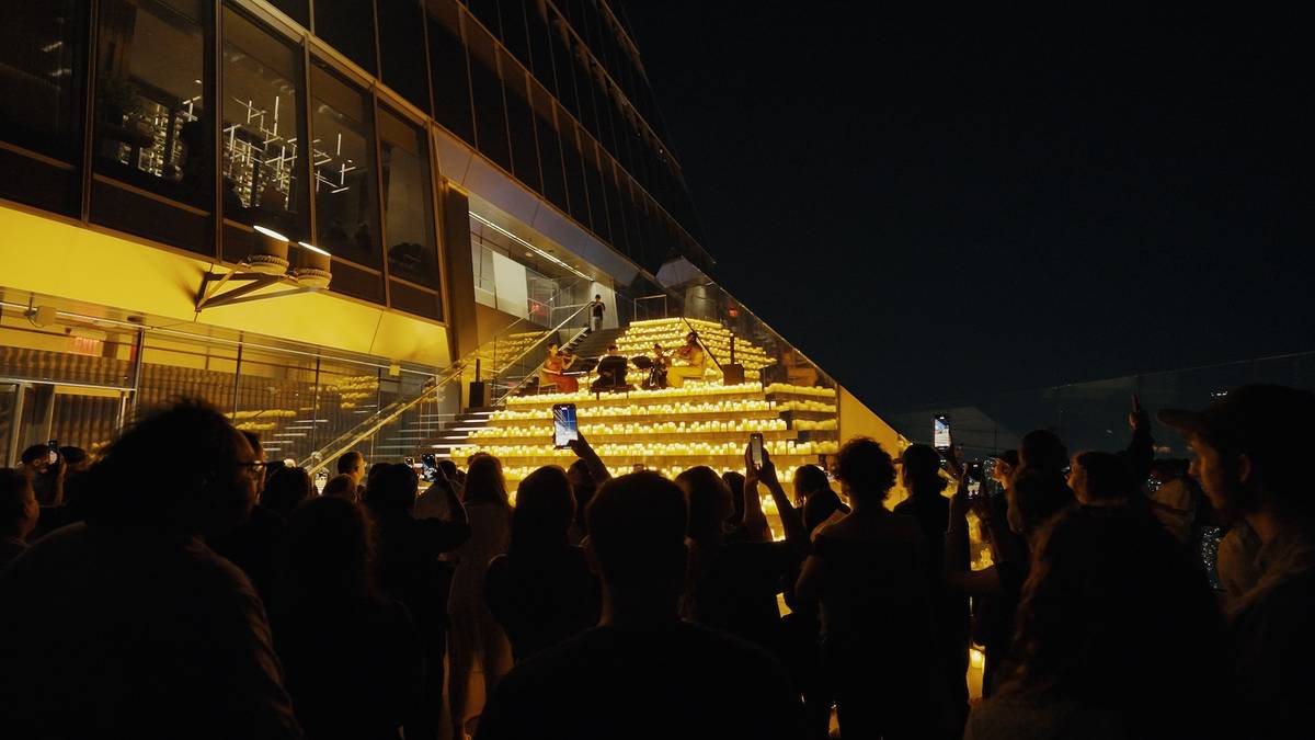 A string quartet plays on a candlelit staircase at Edge with a crowd of people watching, the view of the NYC skyline is reflected on the windows next to them.