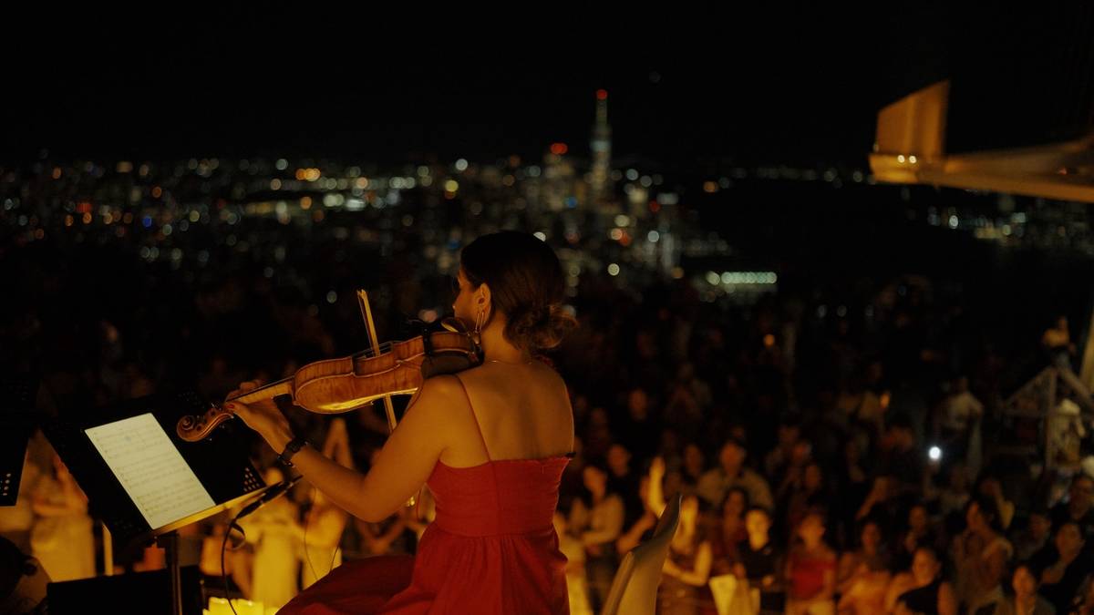 The view from behind a violinist playing to a crowd of people with a distant view of the NYC skyline in the background at Edge.