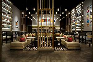 A large couch with pillows, individual tables with round ottomans and tall bar stools, under hanging lights with eclectic pieces of art in front of the fully stocked bar of the Aldo Sohm Wine Bar, NYC