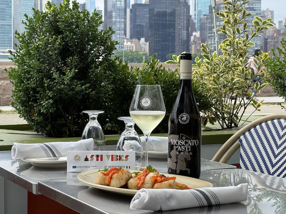 A bottle of Moscato d'Asti wine sits next to a filled class on an outdoor table as Osteria Brooklyn @ LIC alongside a dish from the restaurant.