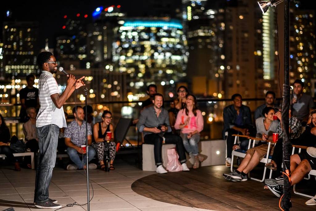 An audience watched a comedian on a roof at night with a lit up city behind.