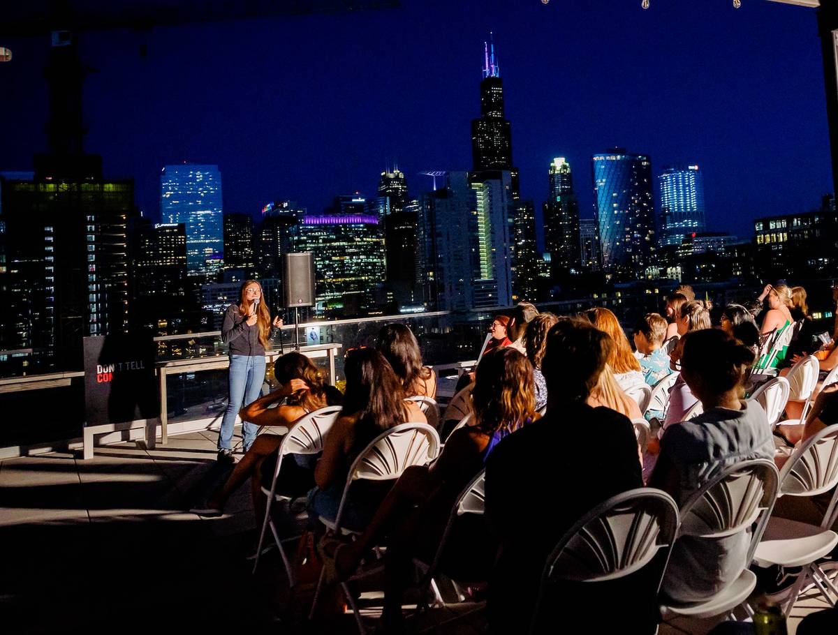 An audience watched a comedian on a roof at night with a lit up city behind.