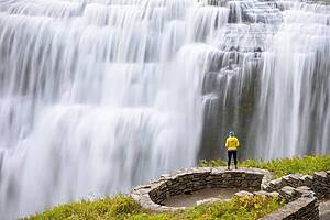 Solo female hiker stands near the edge of a cliff overlooking a giant waterfall. Letchworth State Park, upstate New York, USA