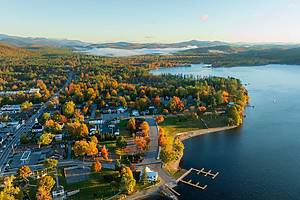 A stunning sunrise over Schroon Lake, NY, with warm light casting a soft glow on the calm water and surrounding autumn-colored forests and mountains.