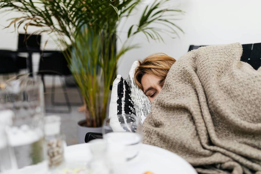 Femme malade sur le canapé avec une couverture sur le visage, des médicaments et de l'eau à proximité.