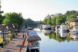 Fairport, NY, USA - July 2nd 2025 - People working and walking along the red brick canal path, in the Village of Fairport. Boats are tied up along the canal, with flowers, American Flags, and a bridge also pictured.