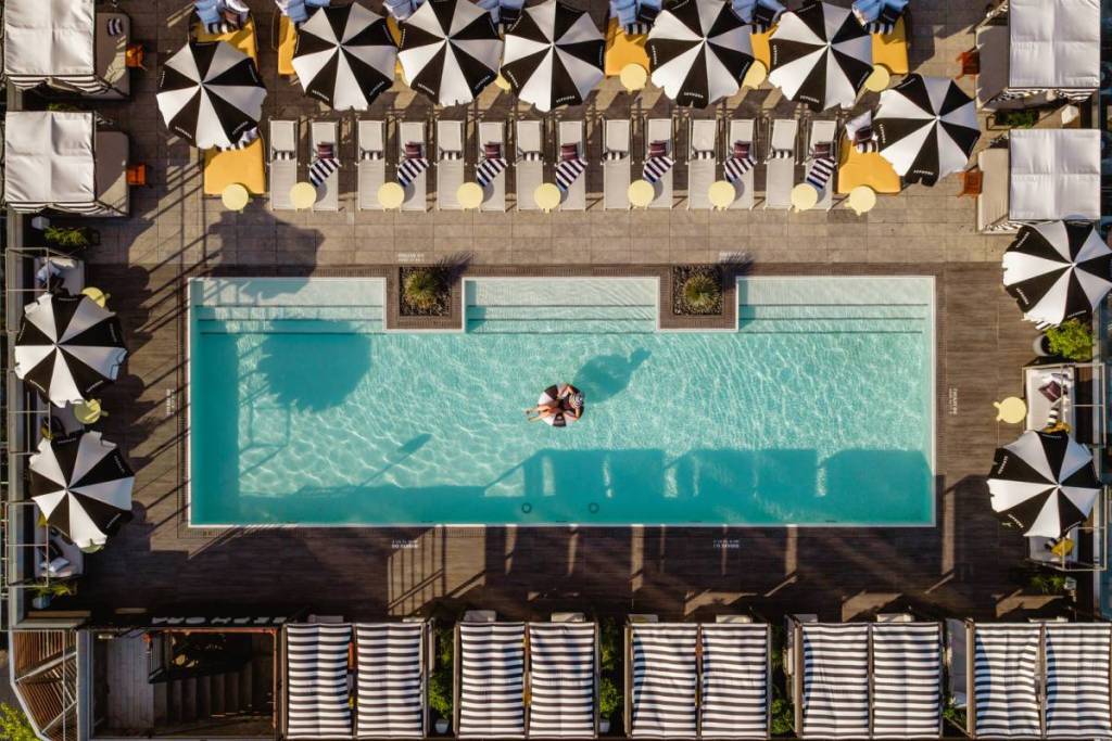 Blick von oben auf eine Person, die den Dachpool im „The William Vale Spa“ in Brooklyn genießt, in einem Schwimmring, umgeben von Sitzgelegenheiten, Liegestühlen und Sonnenschirmen.