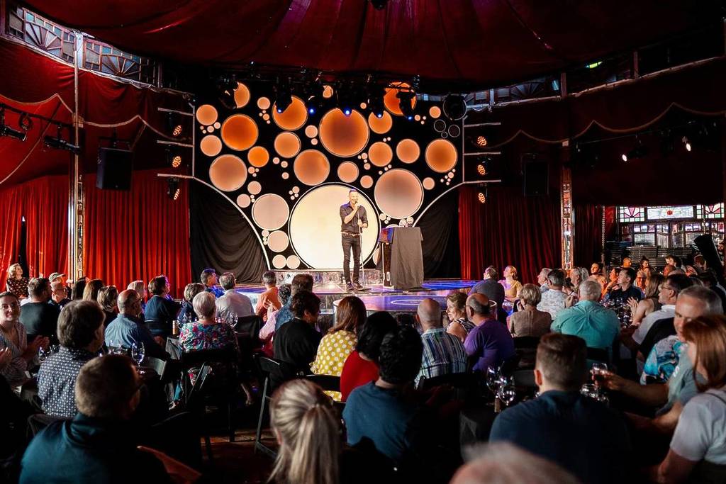 The comedian telling jokes on stage at a comedy show while the audience laugh and drink wine
