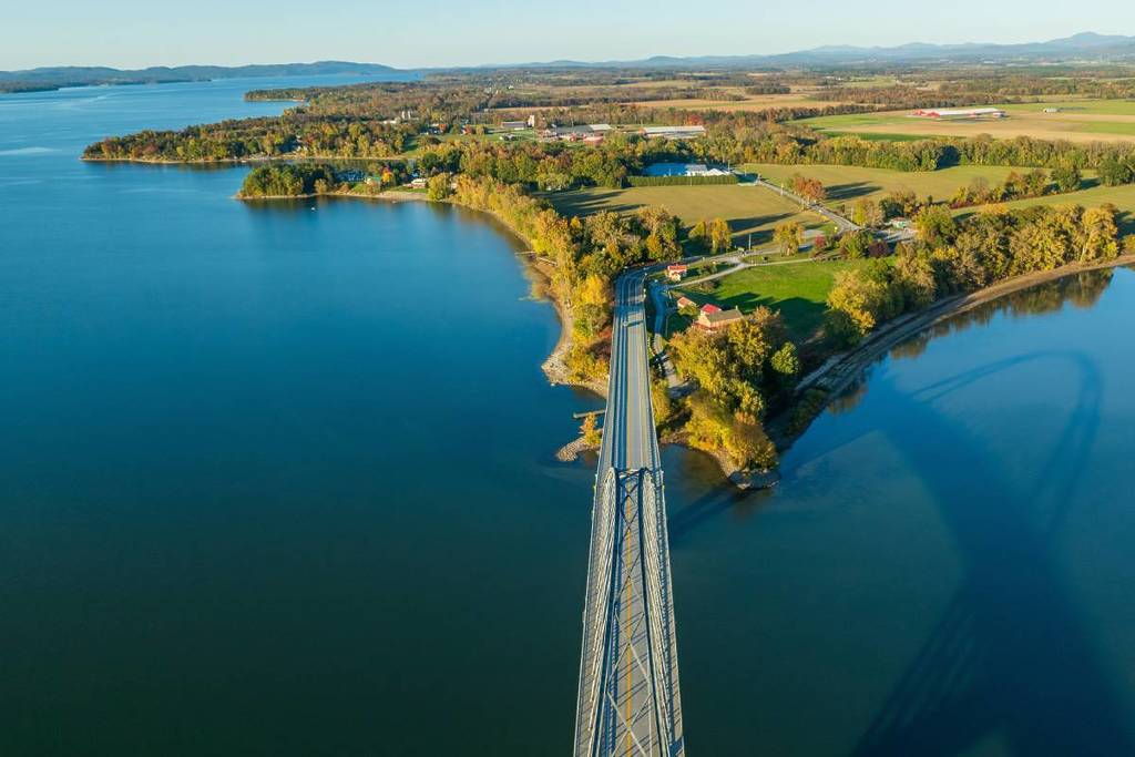 Aerial view of the Lake Champlain Bridge connecting Vermont to New York, spanning calm blue waters with scenic farmland and vibrant autumn trees lining the shoreline
