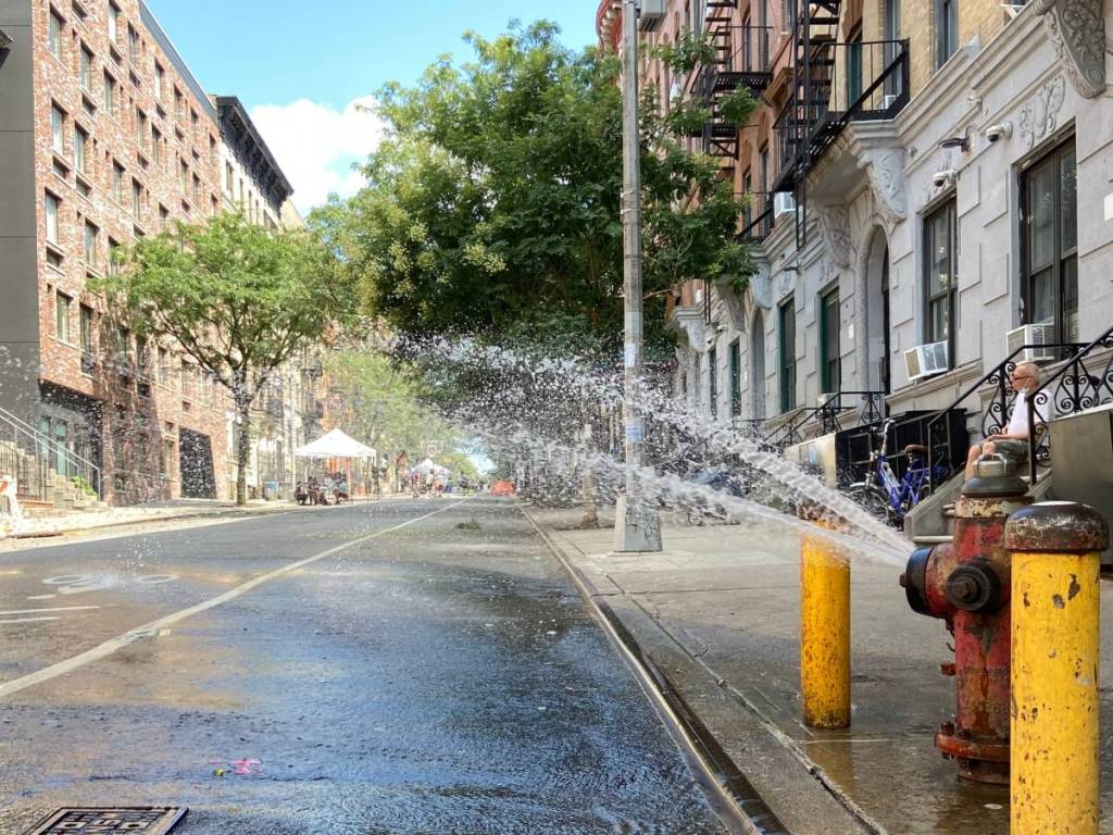 Ein Mann sitzt im Sommer in Washington Heights, New York City, auf der Treppe eines Wohnhauses vor einer verkehrsfreien „Open Street“ mit einem offenen Hydranten