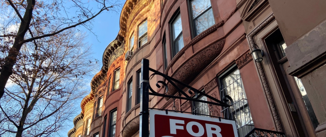 For sale by owner sign hanging in front of a home on a row of historic Harlem brownstones in New York City
