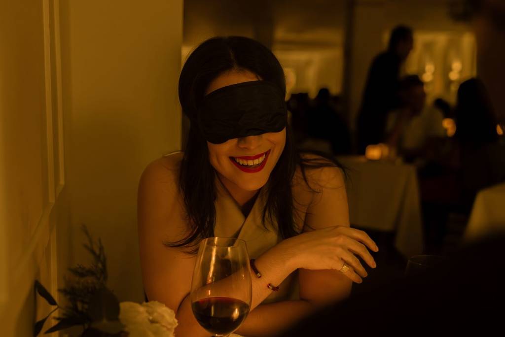A woman wearing a blindfold is smiling at Dining in the Dark.