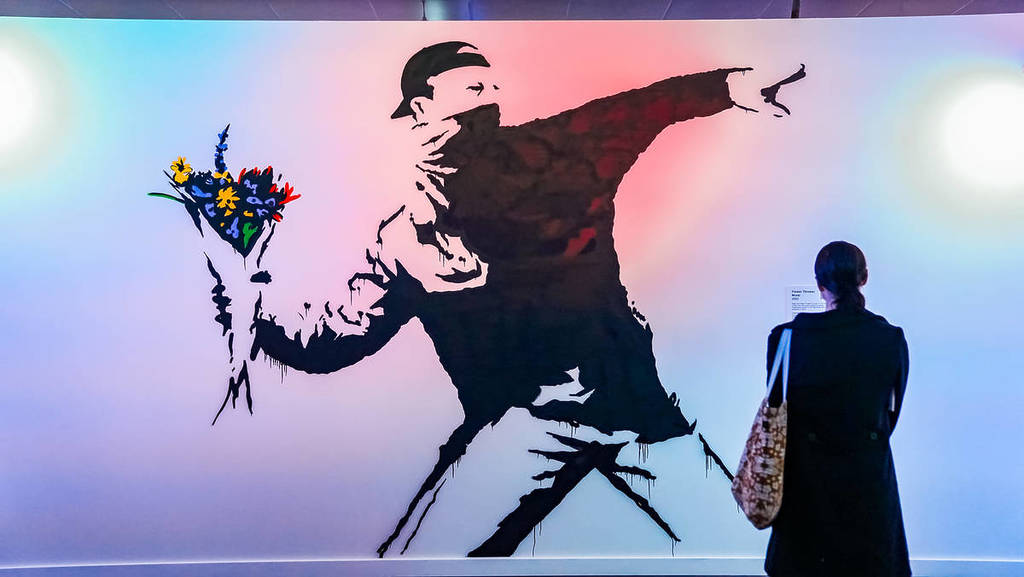 A woman stands in front of a blown-up recreation of The Flower Thrower on a wall at The Art of Banksy.