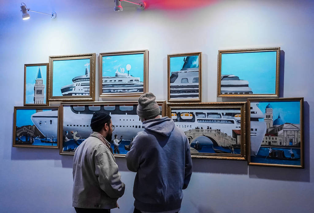 Two men stand in front of multiple works hung on a wall that combine to pay tribute to Banksy's work with the Louise Michel.