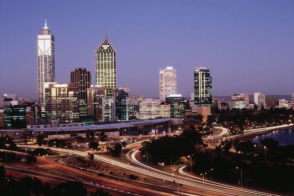 A view of the Perth skyline at dusk with a light purple sky. It features a couple of skyscrapers, the freeway, and a bit of the sea to the right.