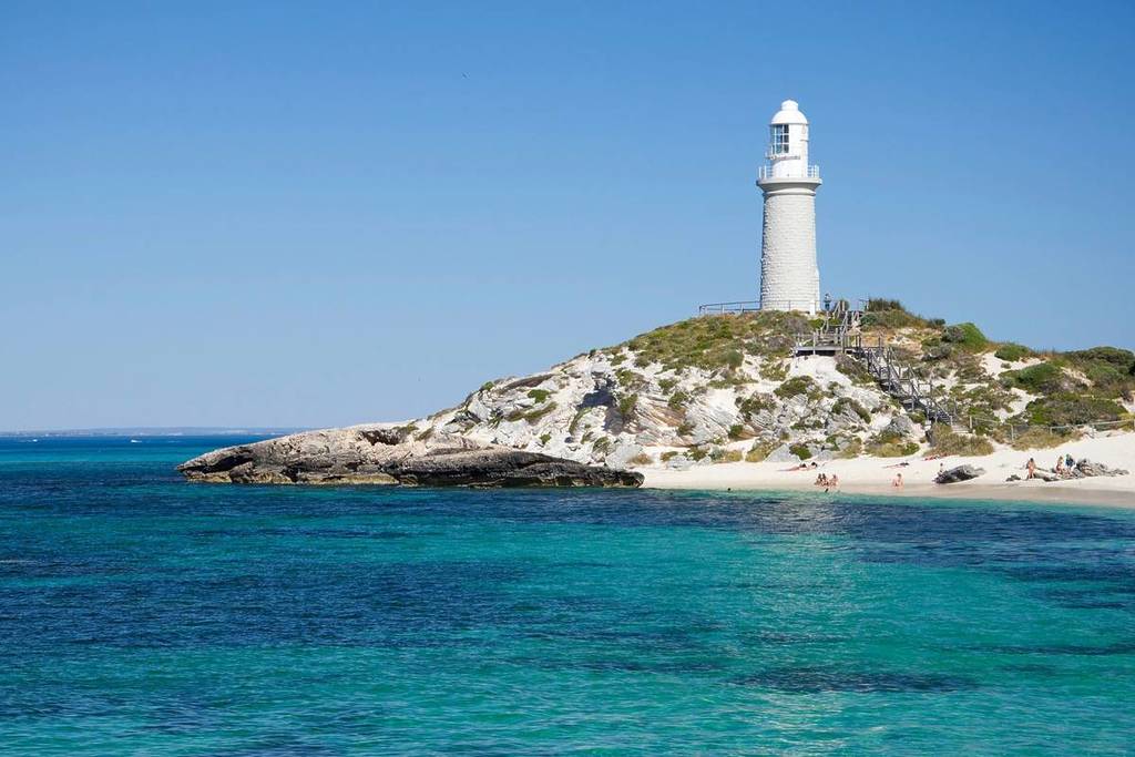 The image shows a white lighthouse in the distance atop a slightly raised piece of land. It is surrounded by clear, blue water on three sides with corals peeking underneath.
