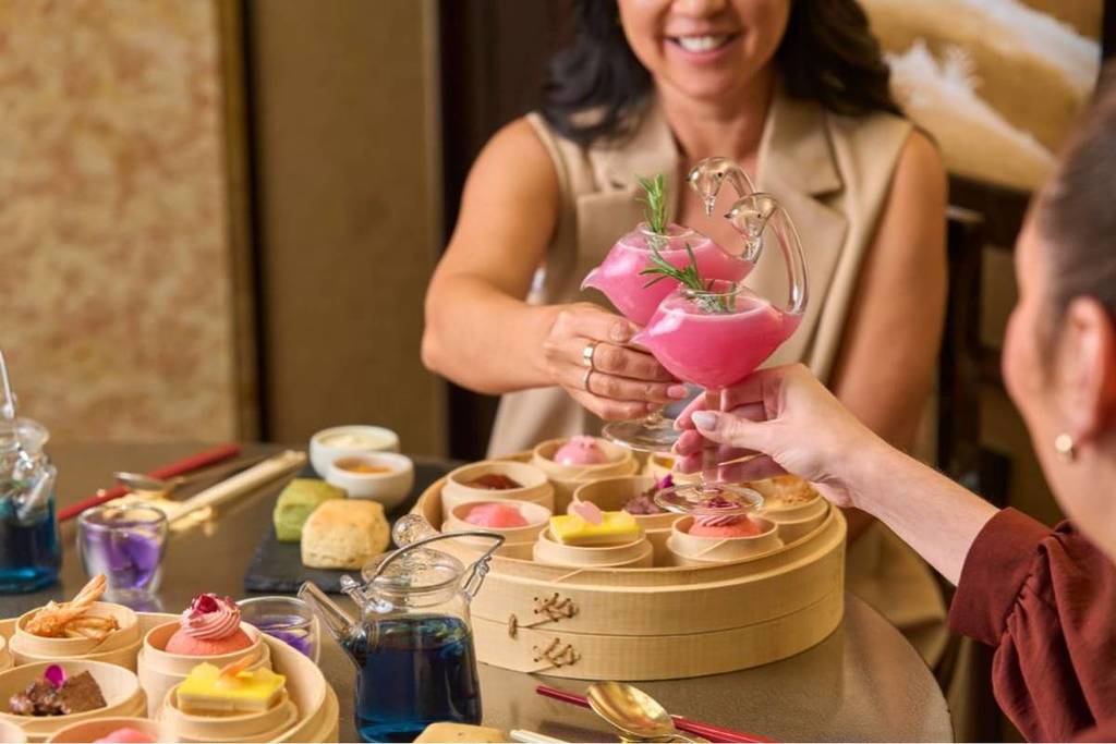The image shows two women toasting wine glasses filled with a pink substance while surrounded by bamboo baskets of dimsum as part of the high tea at Silks, Crown Perth for Mother's Day.