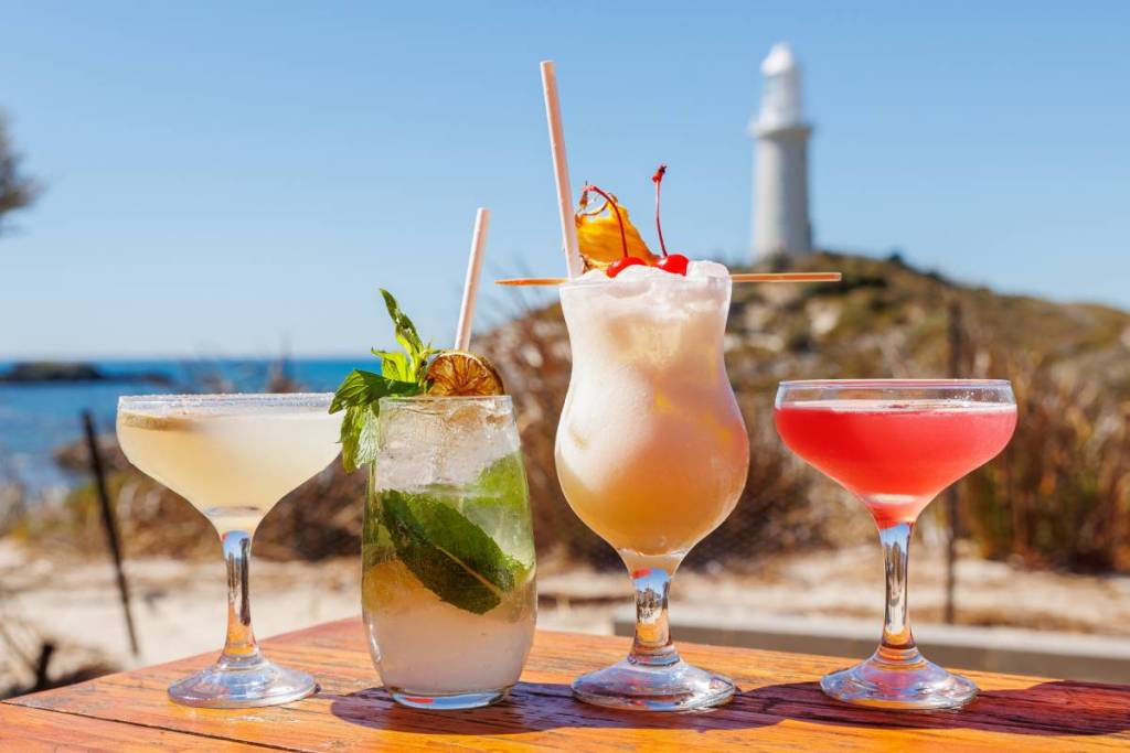 A bunch of colourful drinks on a table against the beach with a lighthouse visible behind as well.