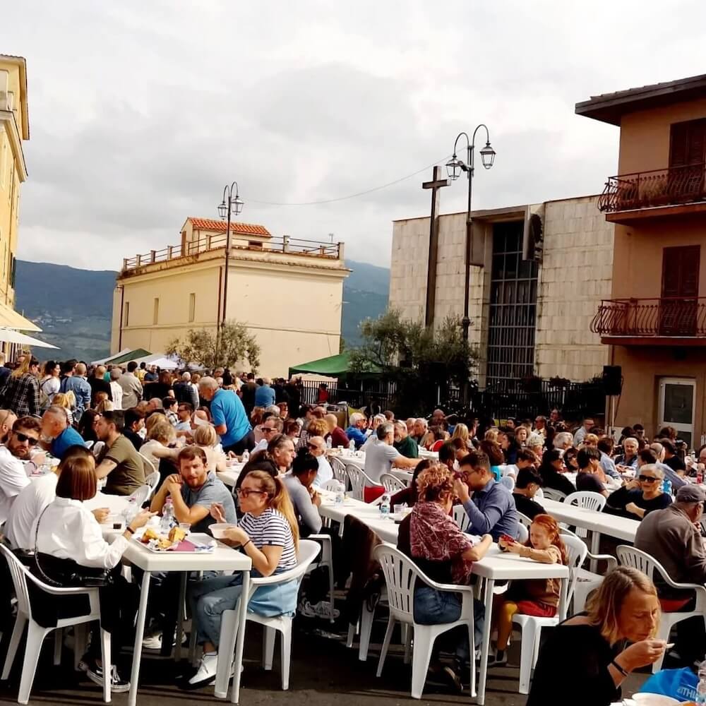Dorffest in Nerola: Eine Menschenmenge sitzt an langen weißen Tischen im Freien und isst genüsslich. Im Hintergrund die historischen und modernen Gebäude des Dorfes, darunter eine Fassade mit einem großen Kreuz, und die umliegenden Hügel unter einem bewölkten Himmel.