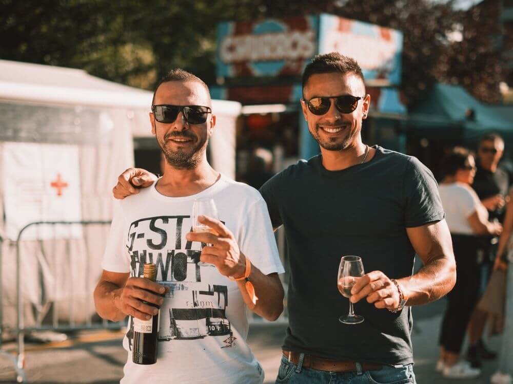 Zwei lächelnde Männer mit Sonnenbrillen im Vordergrund, die bei einem lebhaften Open-Air-Fest mit Weingläsern anstoßen.