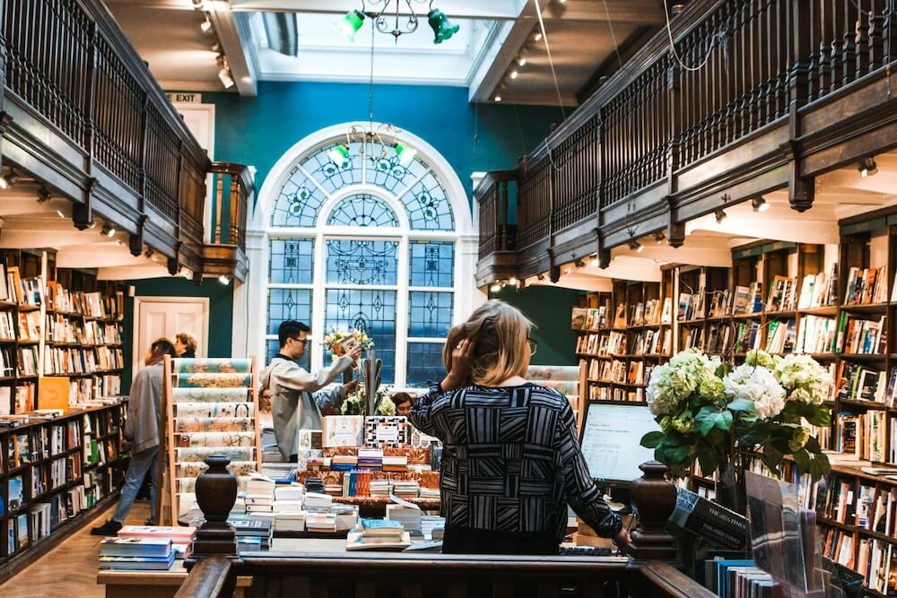 Interno di una libreria storica a due piani con mezzanini in legno e una grande finestra a vetrata colorata. In primo piano, una donna bionda guarda lo schermo di un computer accanto a un mazzo di ortensie bianche. Altri clienti guardano i libri sugli scaffali.
