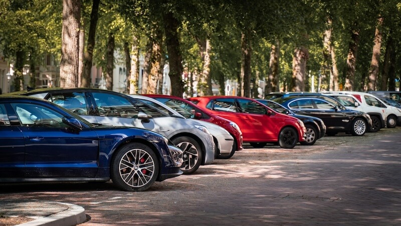 Fila di auto parcheggiate in un parcheggio lungo un viale alberato, con veicoli disposti in sosta su entrambi i lati della strada.