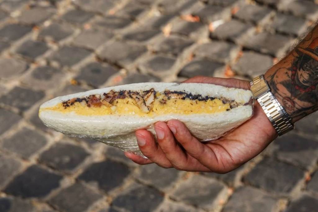 Tramezzino gourmet farcito tenuto da una mano con orologio e tatuaggio su uno sfondo di sampietrini romani.