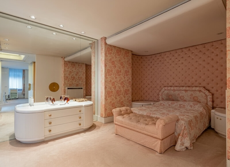 Camera da letto di Raffella Carra, elegante dai toni rosa e crema, con letto matrimoniale, panca ai piedi del letto, carta da parati floreale e comò bianco davanti a una pareti di specchi.