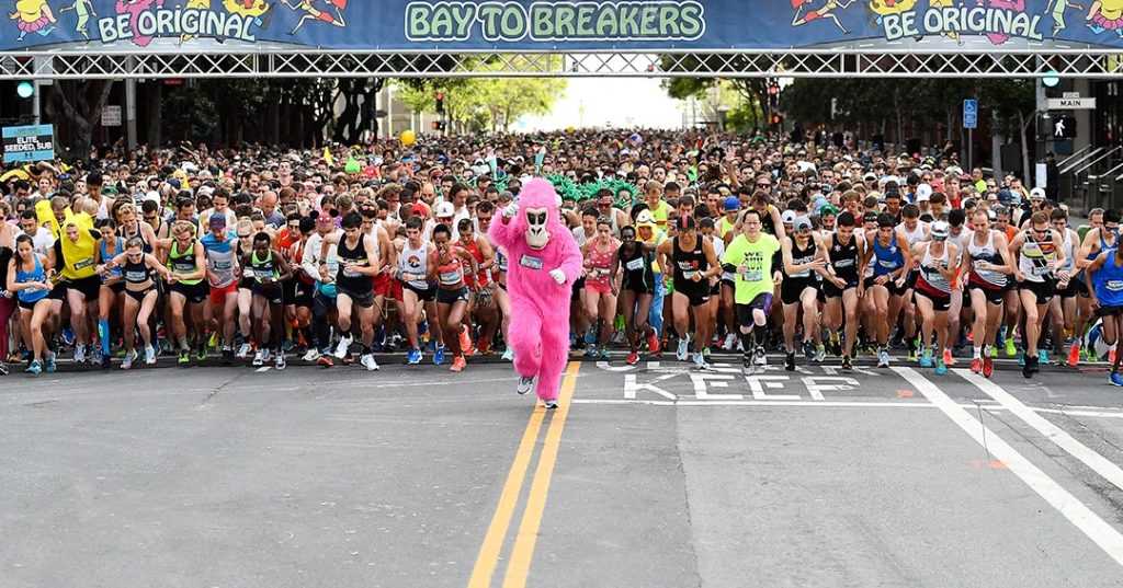 Hundreds of runners take off at the start line of Bay to Breakers, led by a person in a pink monkey suit.