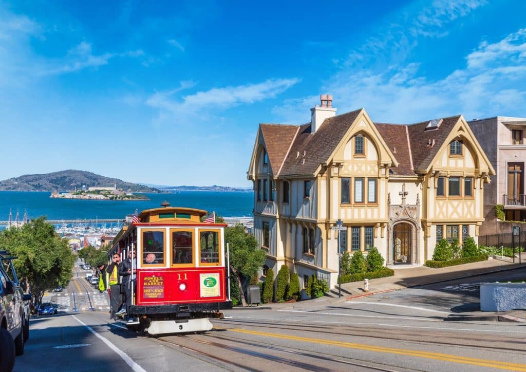 Red cable car coming up Hyde Street with Alcatraz in the background on a sunny day in San Francisco