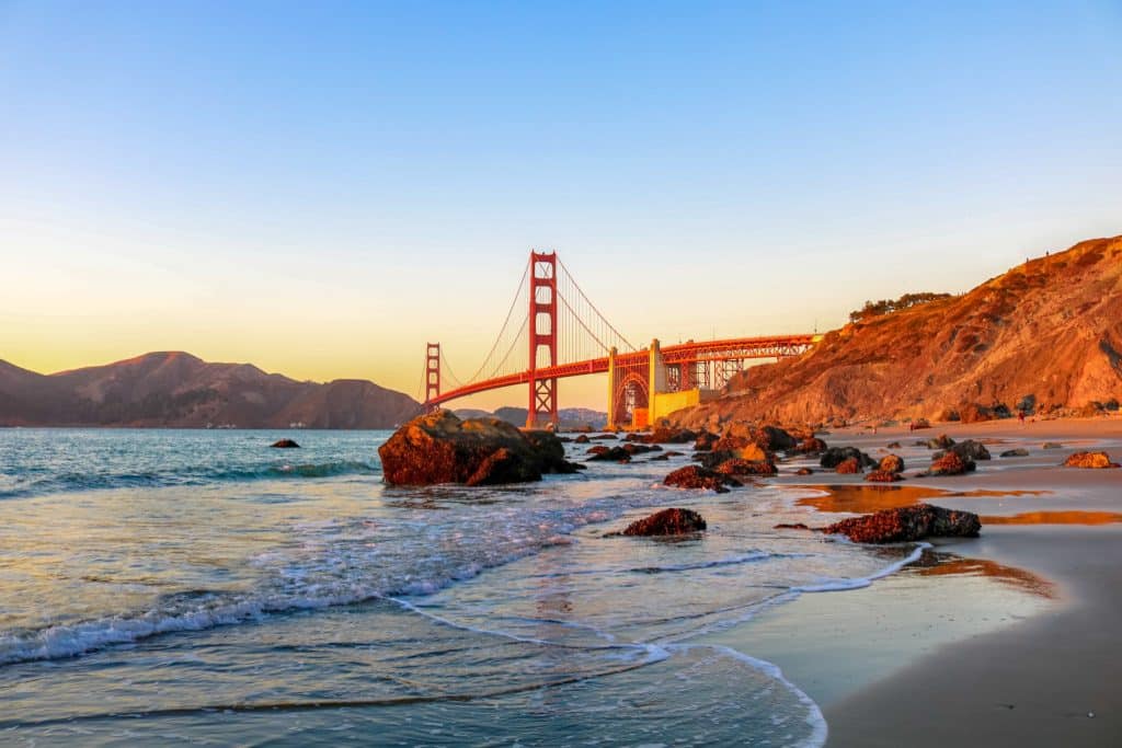 Marshall's Beach at golden hour with. the waves crashing against rocks and the Golden Gate Bridge in the Distance.