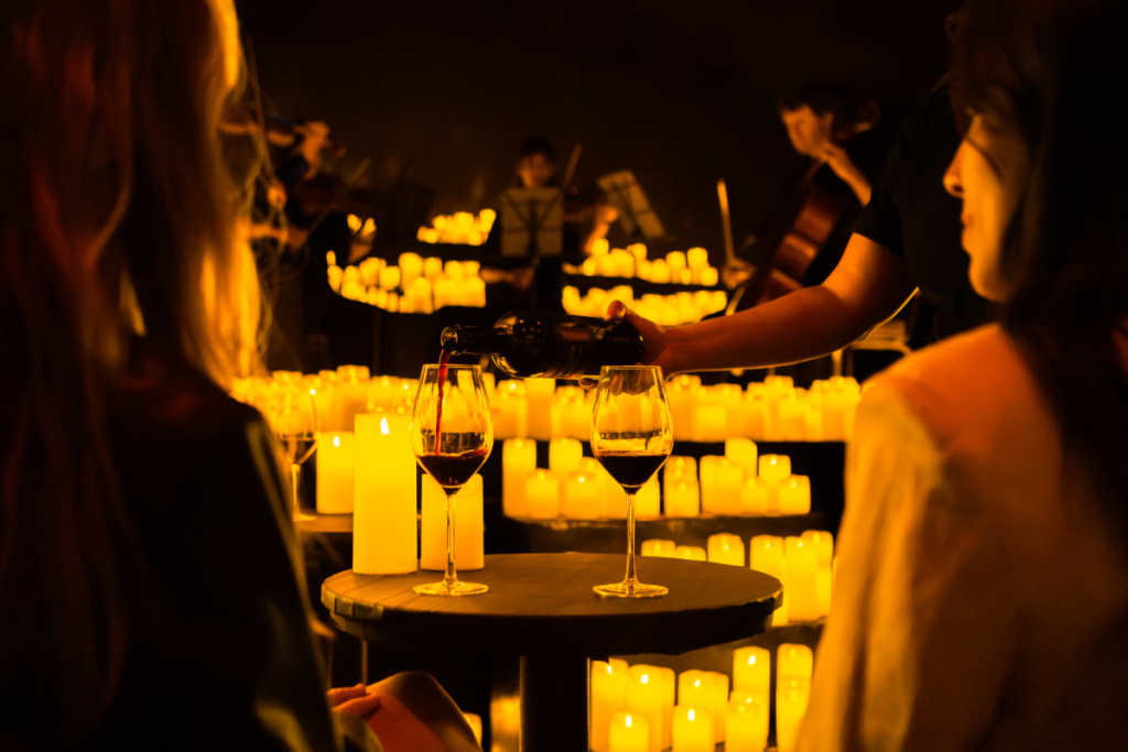 Dos mujeres sentadas en una mesa con dos copas de vino tinto, contemplando la actuación de un cuarteto de cuerda en medio de un mar de velas