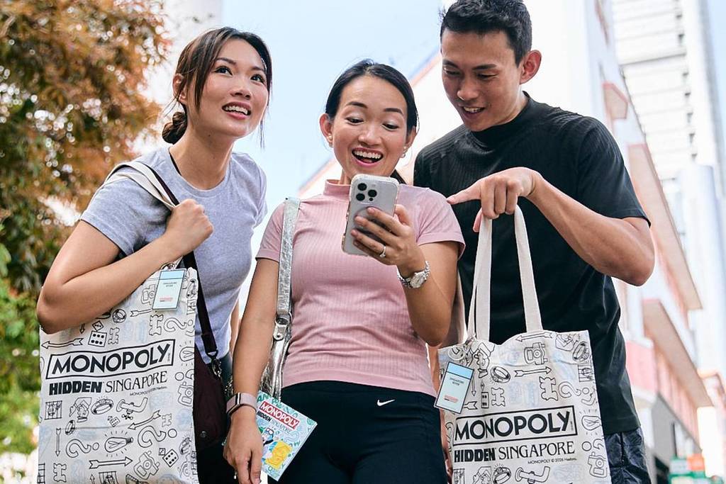 A trio of explorers solving clues via WhatsApp with their exclusive MONOPOLY: HIDDEN - Singapore River tote bags