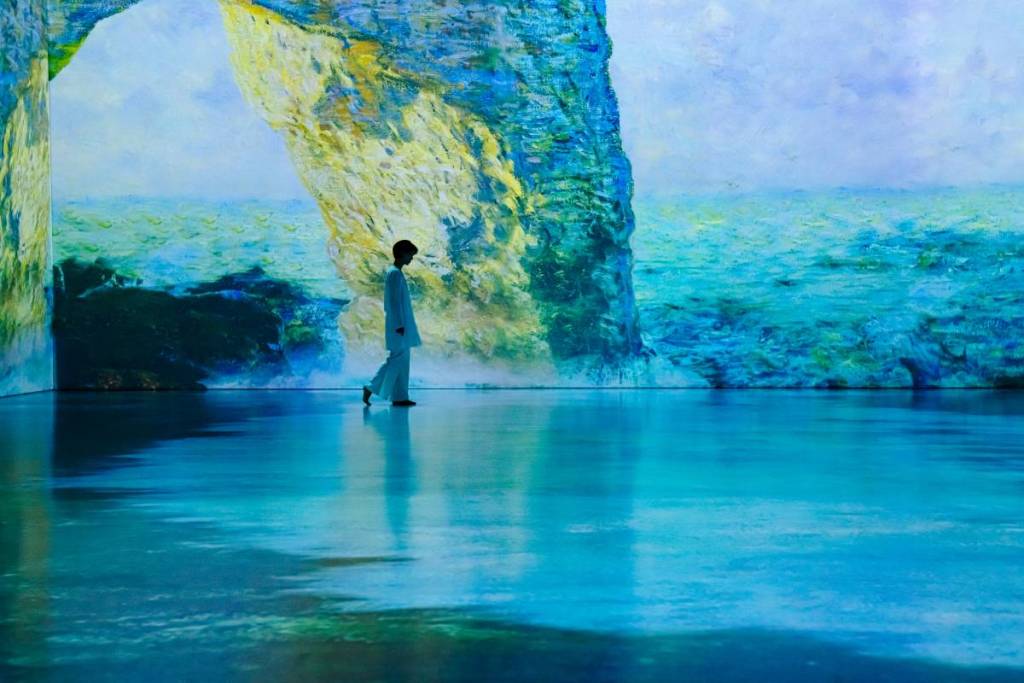 A woman walking along a projected scene of The Manneporte at Monet Inside