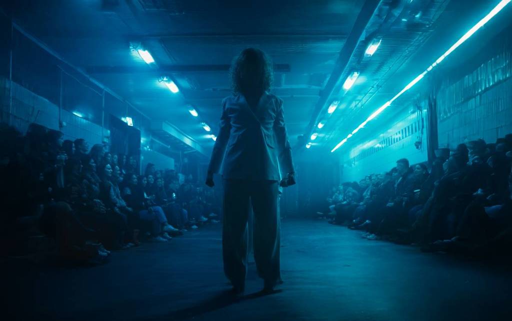A dancer performing on stage in white shirt and trousers in front of the crowd sitting closely in a dim-lit blue light