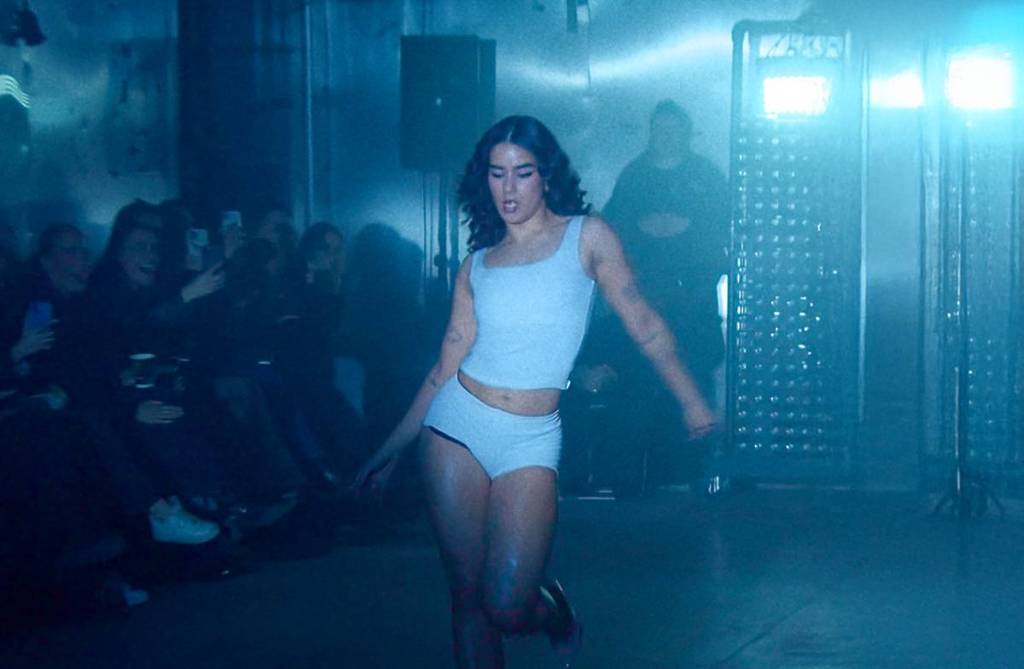 A female dancer performing on stage in white shorts and top in front of the crowd sitting closely in a dim lighting