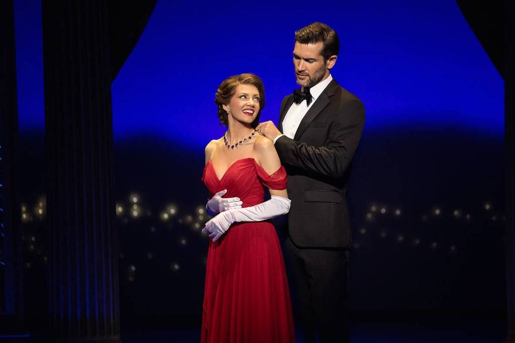 A man in a tuxedo places a necklace on a woman in a red gown on stage for the Pretty Woman Musical in Sydney